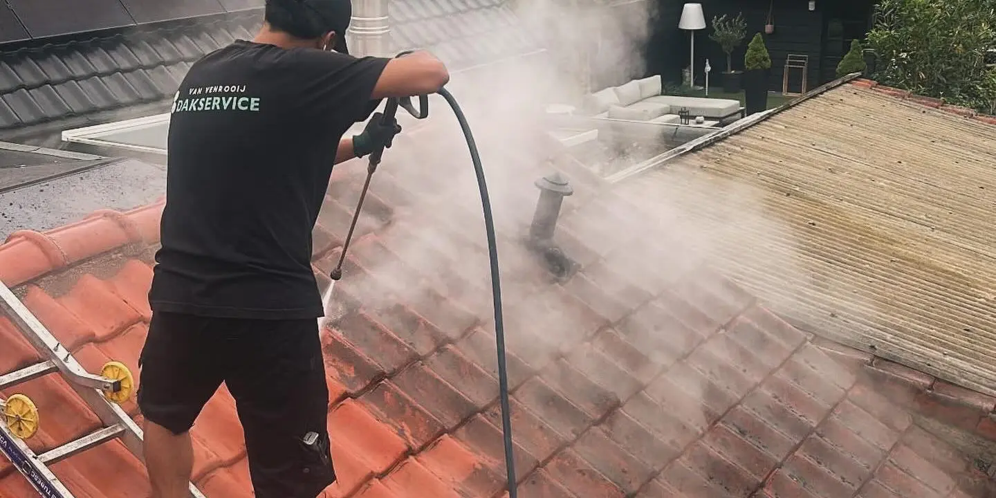 Dakdekker met Van Venrooij Dakservice shirt reinigt dakpannen met hogedrukreiniger vanaf ladder op hellend dak.