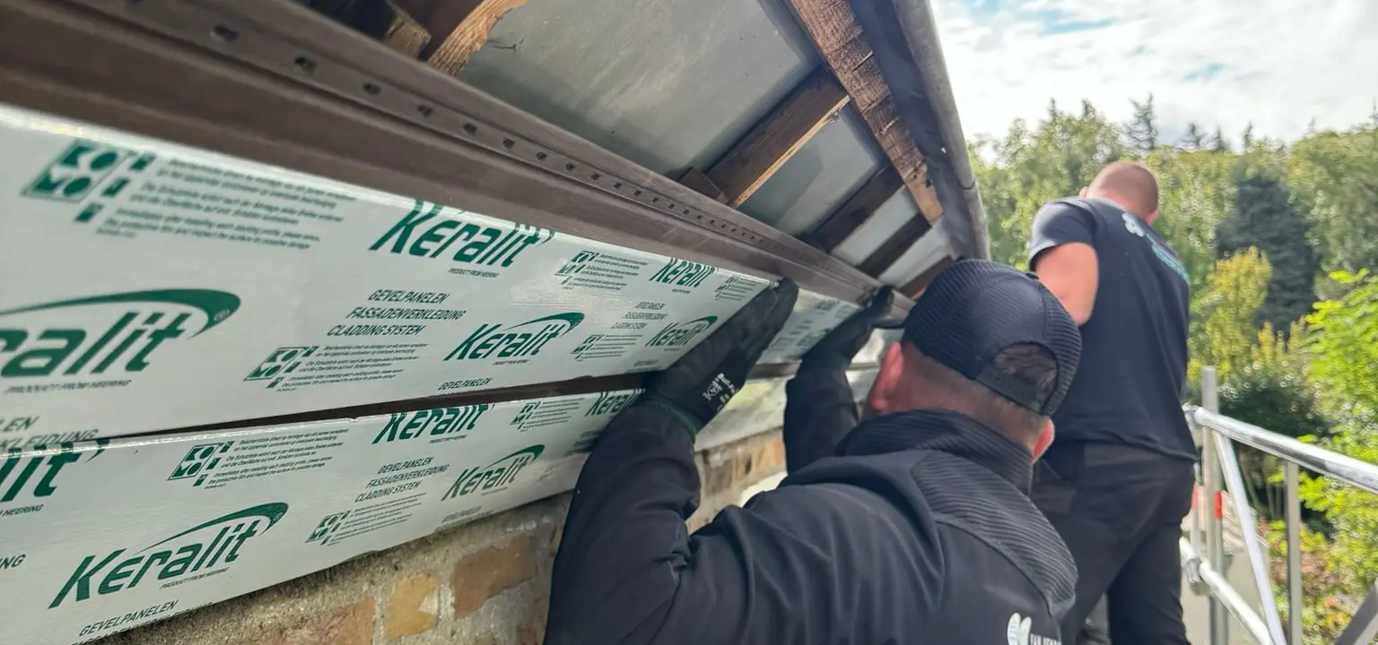 Dakdekkers plaatsen isolatiepanelen onder dakrand van een woning, werken op steiger bij zonnige lucht.
