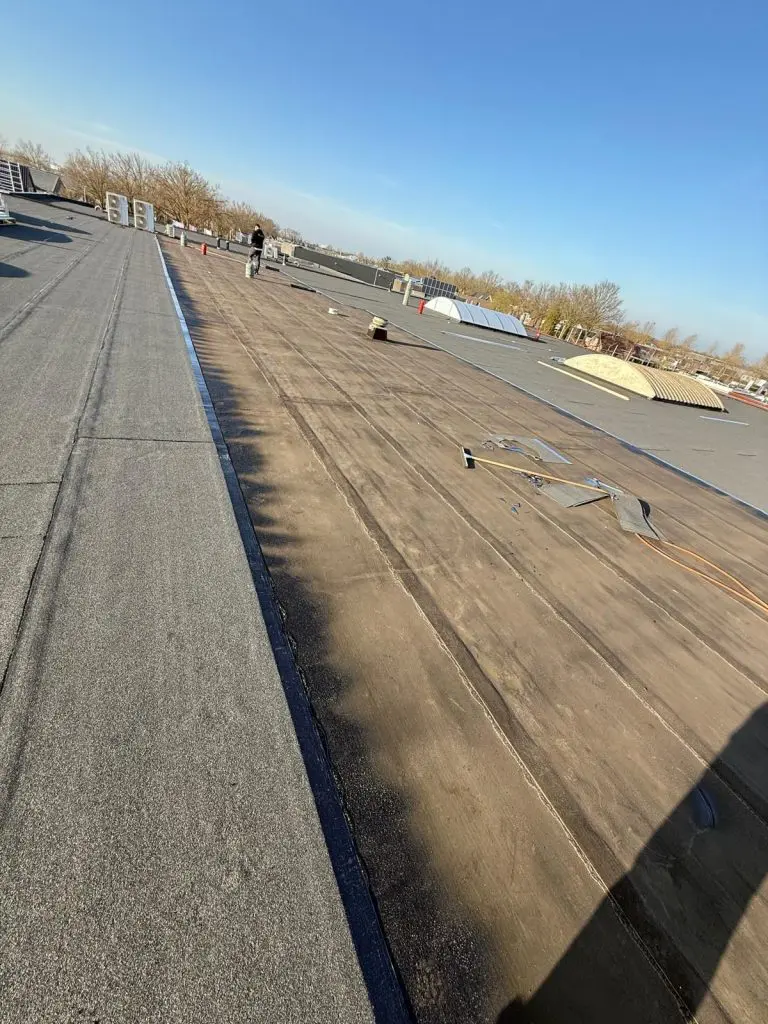 Dakdekker werkt op plat dak met bitumen dakbedekking onder heldere blauwe lucht en gereedschap verspreid.