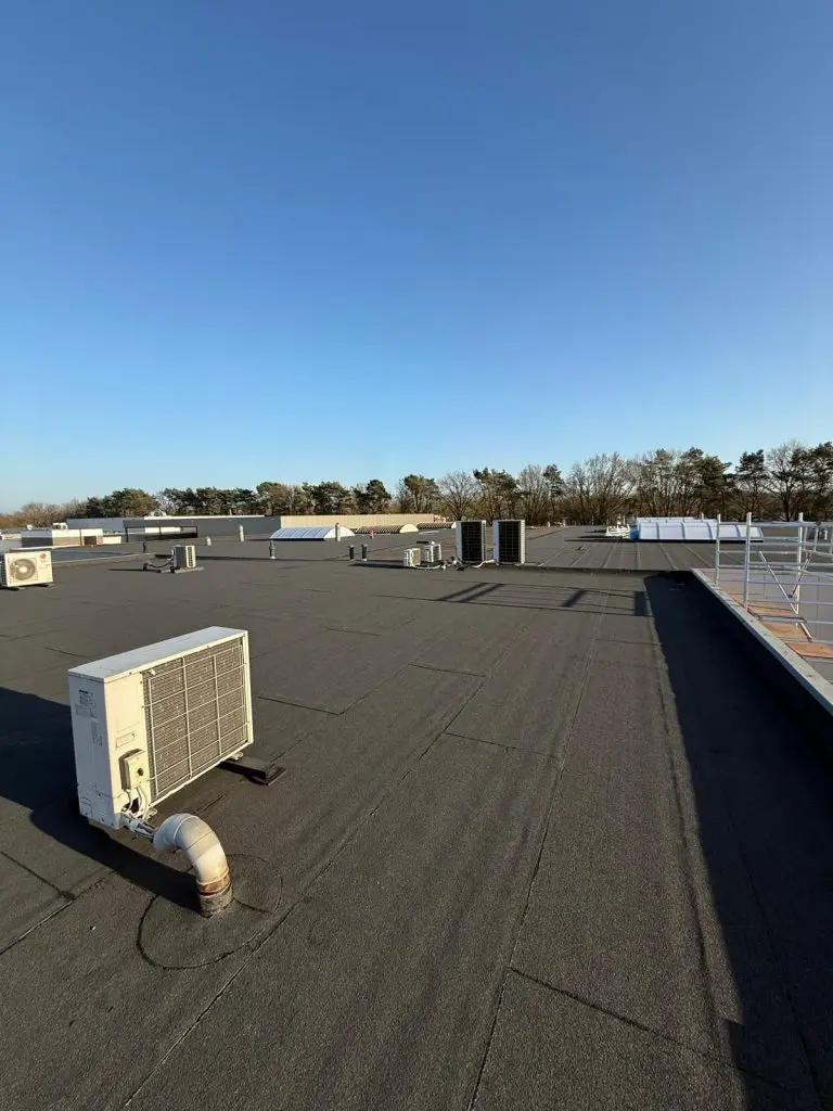 Plat dak met bitumenbedekking en meerdere airconditioningunits onder een heldere blauwe lucht.
