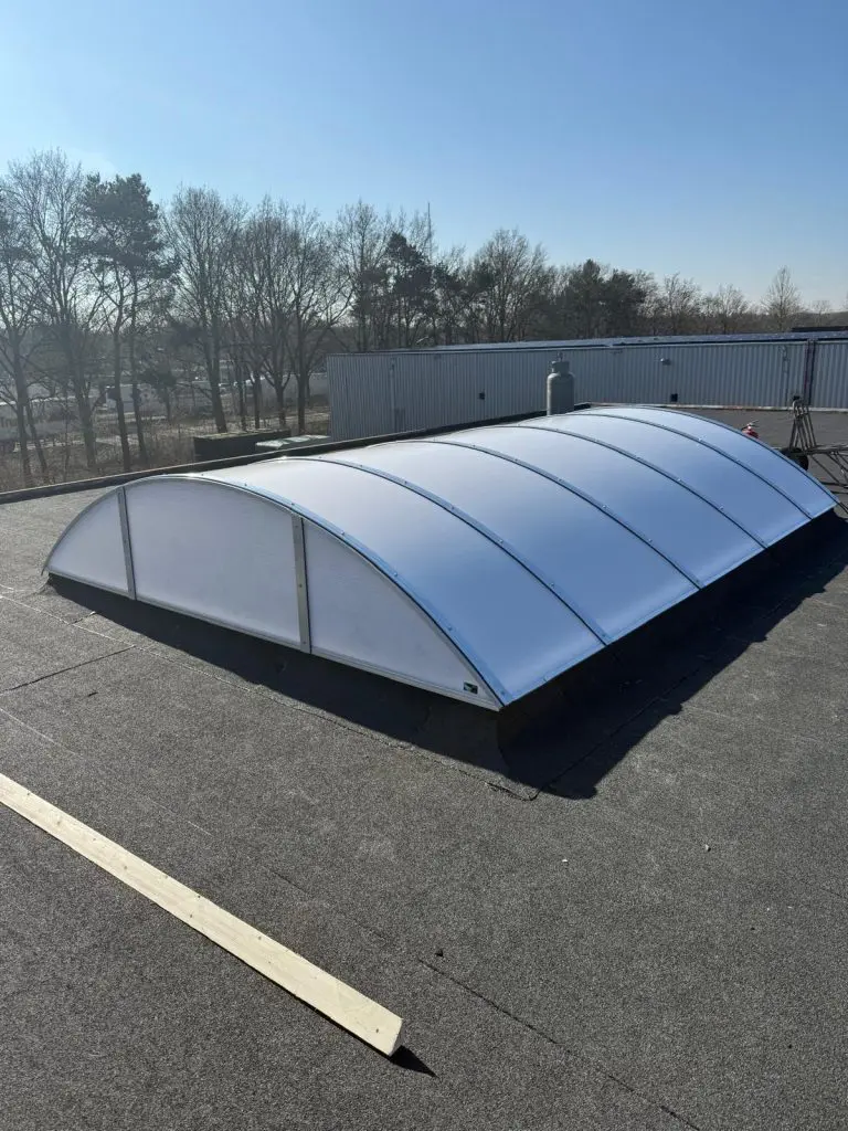 Plat dak met zwarte bitumenbedekking en een gebogen lichtkoepel van kunststofpanelen onder blauwe lucht