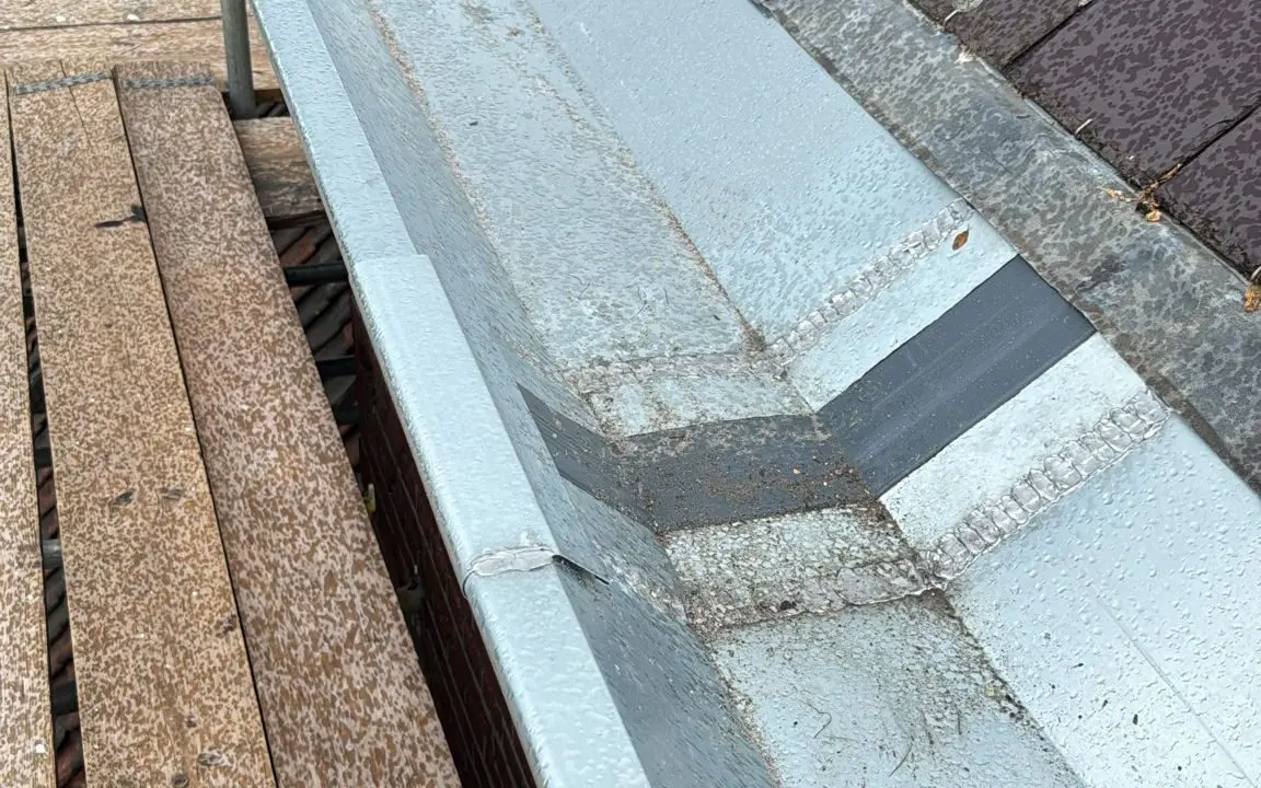Dakrand met metalen dakgoot en bruine dakpannen, steigerplanken zichtbaar voor dakwerkzaamheden.