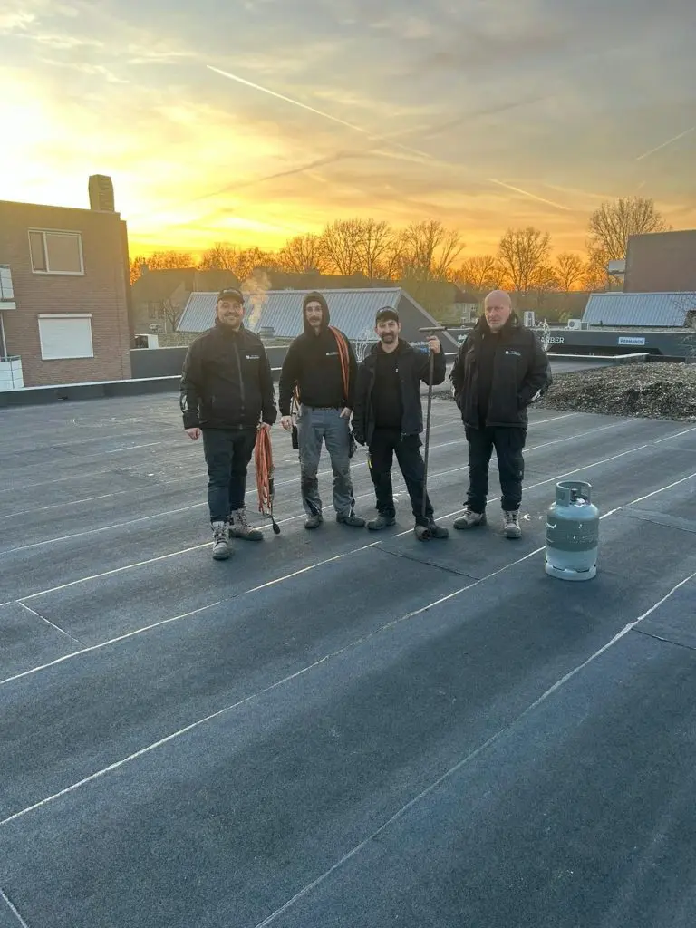 Vier dakdekkers staan op een plat dak met bitumen bij zonsondergang, gereedschap en gasfles zichtbaar.