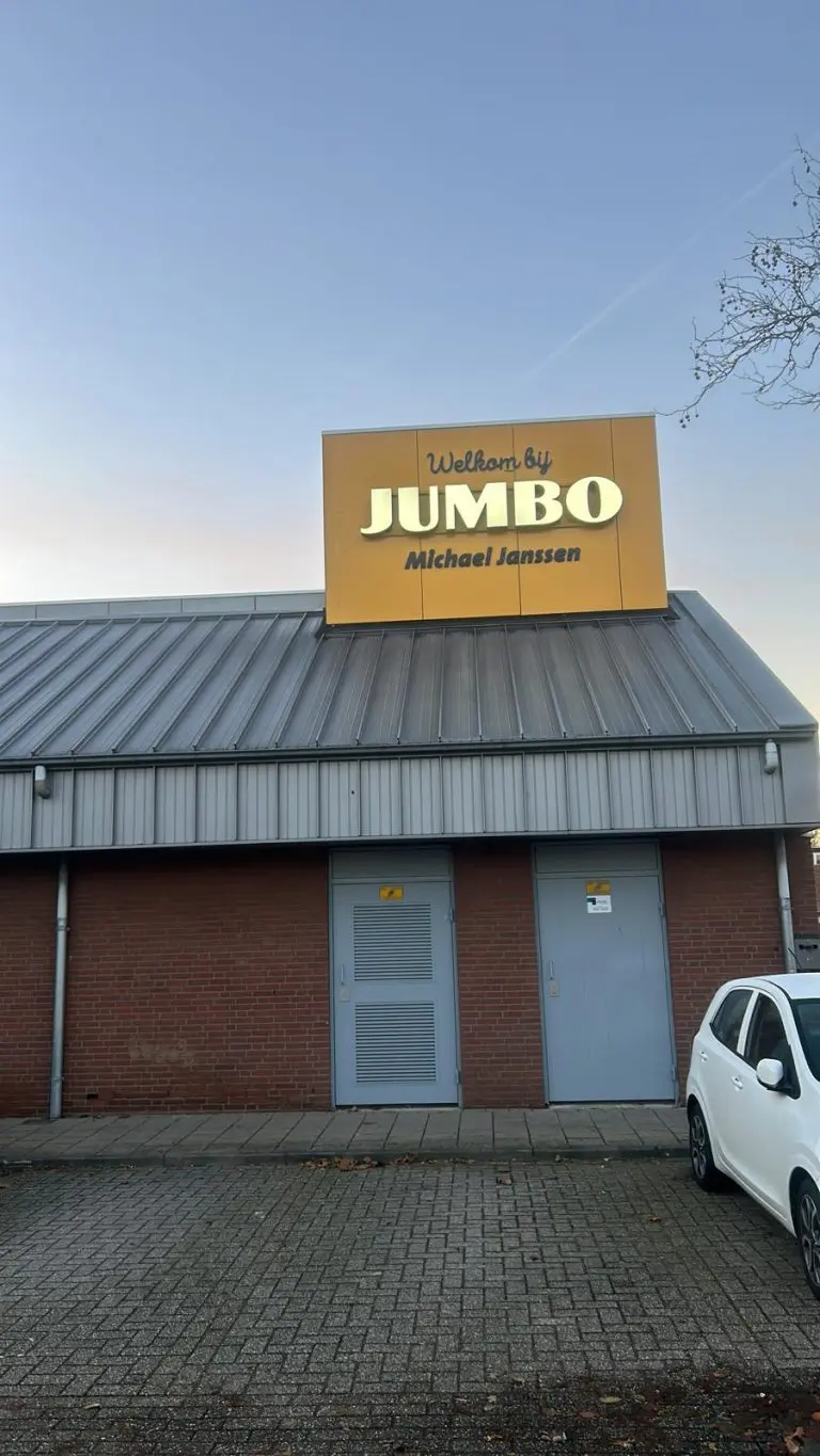 Dak met metalen dakbedekking en twee grijze deuren onder een geel reclamebord met tekst Jumbo Michael Janssen.