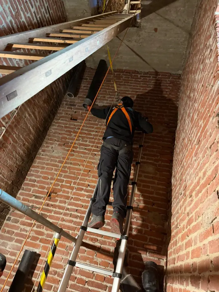 Dakdekker met veiligheidsharnas klimt op uitschuifladder in smalle ruimte met bakstenen muren voor dakwerkzaamheden.