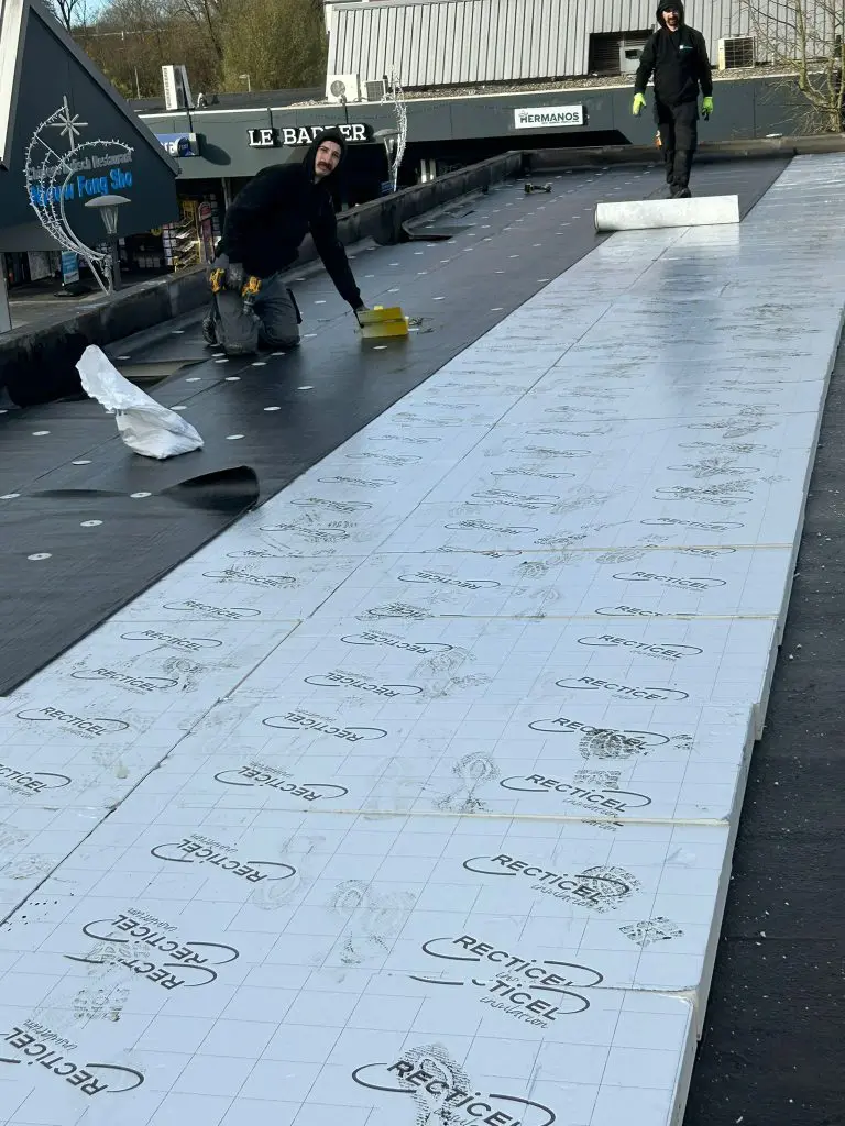 Twee dakdekkers leggen isolatieplaten en bitumen dakbedekking op een plat dak bij een winkelcentrum.