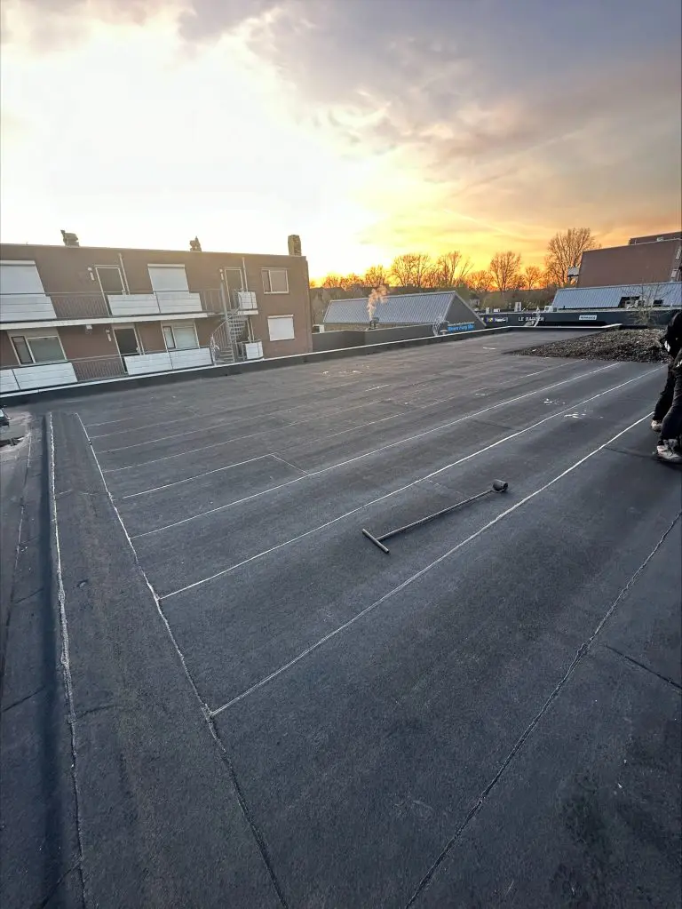 Dakdekker werkt aan het aanbrengen van bitumen dakbedekking op een plat dak bij zonsondergang.