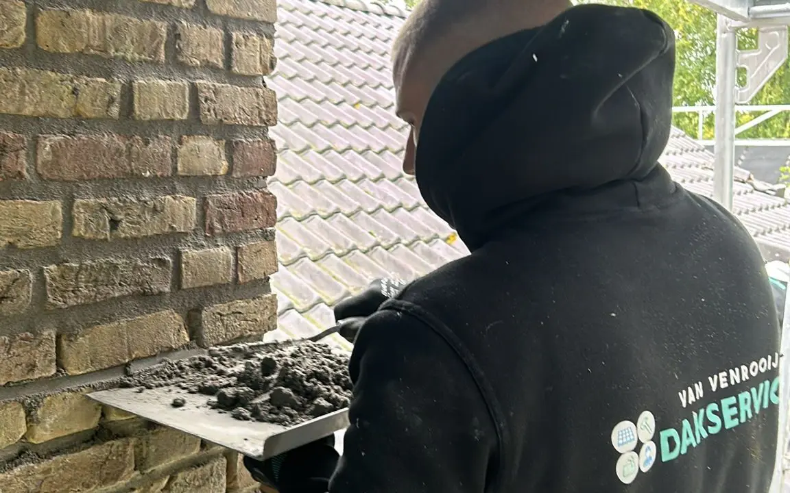 Dakdekker met Van Venrooij Dakservice hoodie werkt aan voegen van een schoorsteen op een steiger bij een pannendak.