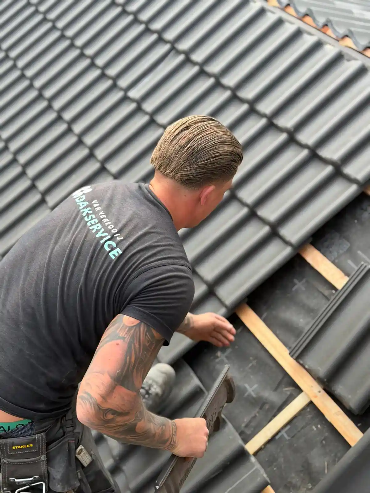Dakdekker met tattoo legt grijze dakpannen op houten latten tijdens dakwerkzaamheden.