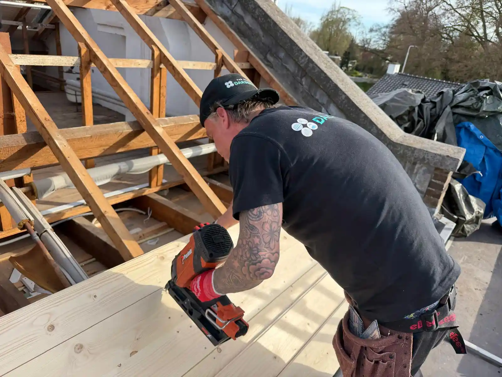 Dakdekker met tattoo spijkert houten planken vast op dakconstructie tijdens dakwerkzaamheden in de zon.