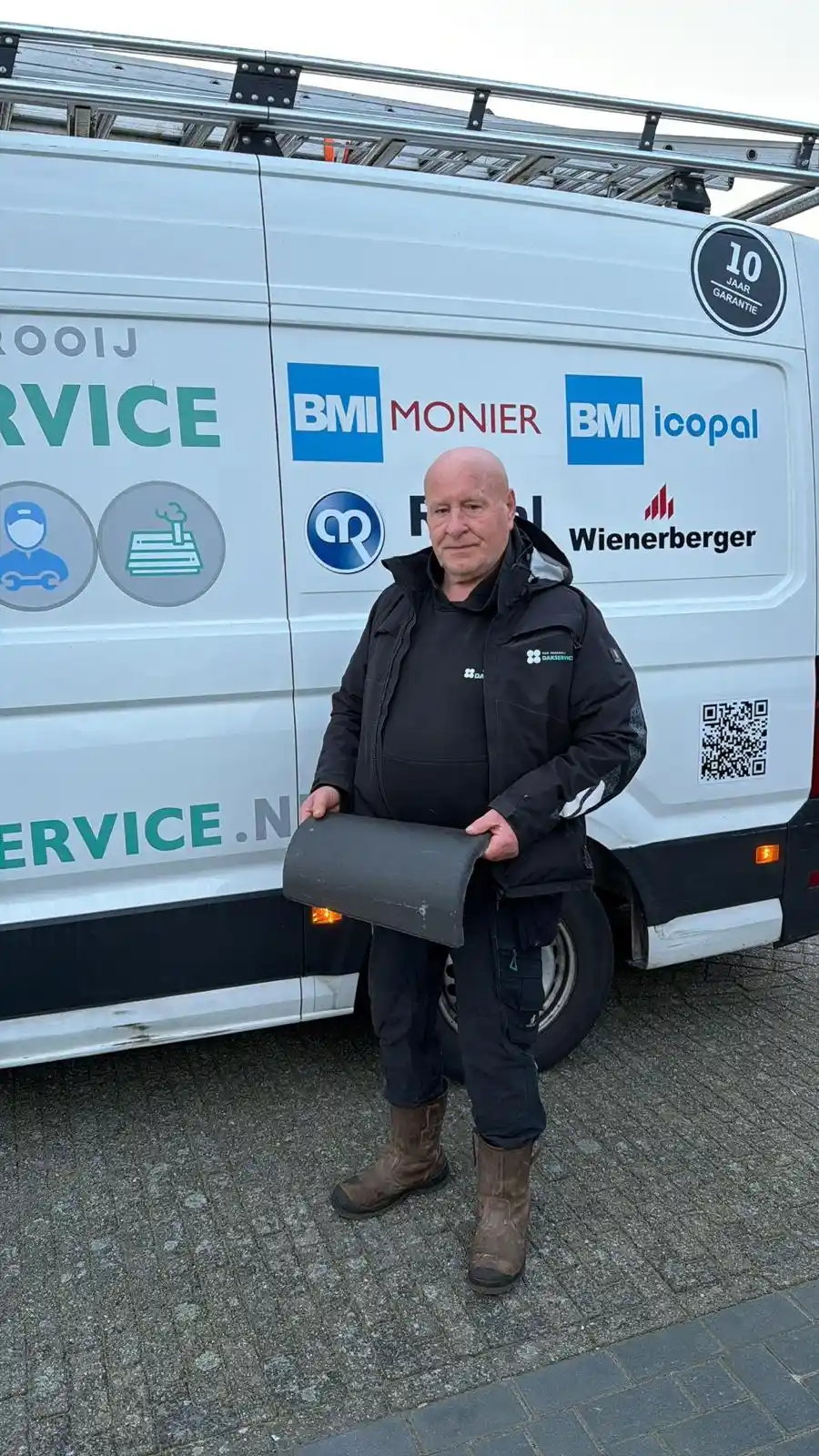 Dakdekker in werkjas met Van Venrooij Dakservice-logo houdt een dakpan vast naast een bedrijfsbus met dakmerken en logo's.