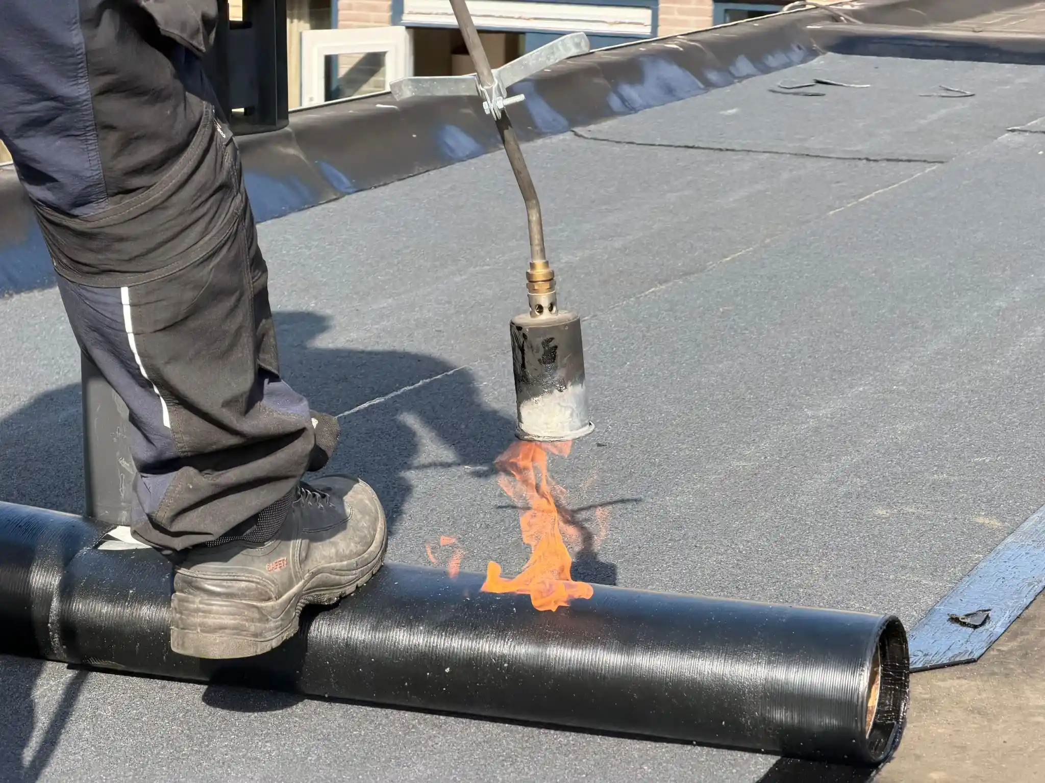 Dakdekker verwarmt bitumen met brander om dakbedekking op plat dak aan te brengen, zicht op werklaarzen en rol bitumen.
