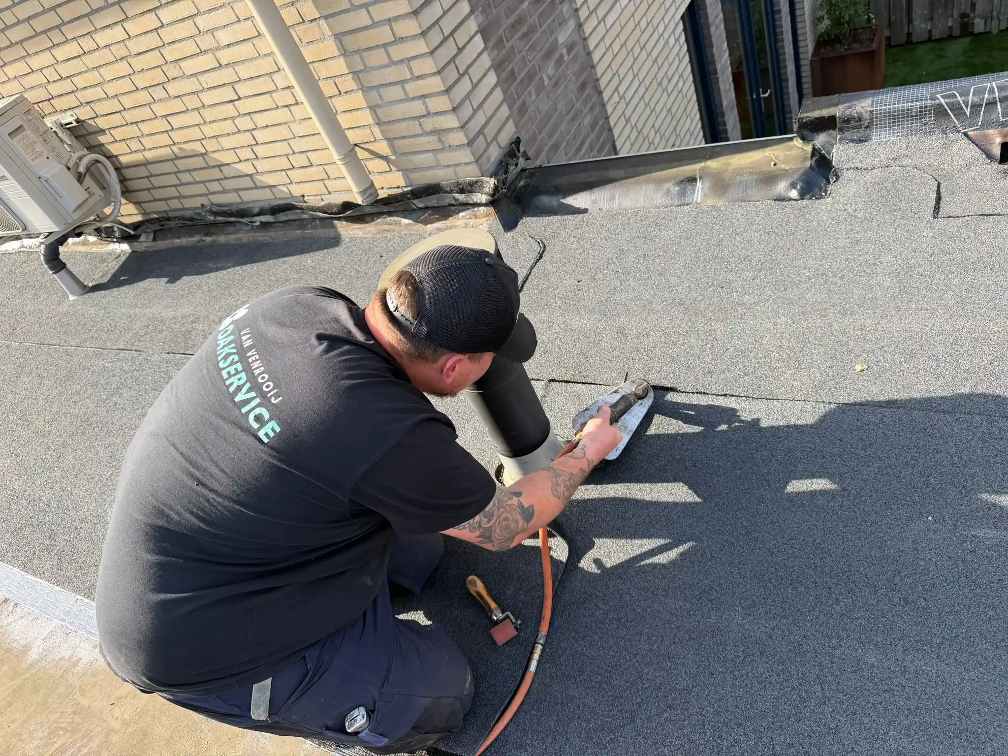 Dakdekker met gereedschap werkt aan bitumen dakbedekking bij dakdoorvoer naast bakstenen gevel.