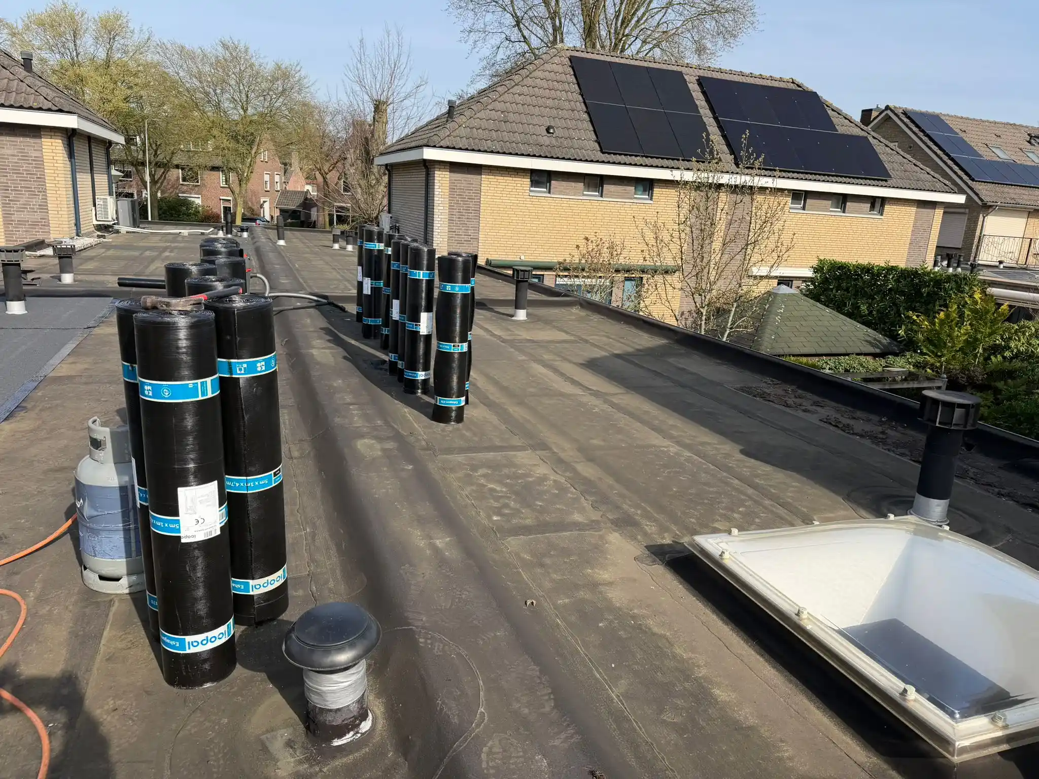 Plat dak met rollen bitumen dakbedekking en zonnepanelen op aangrenzende schuine daken in woonwijk bij zonnig weer.