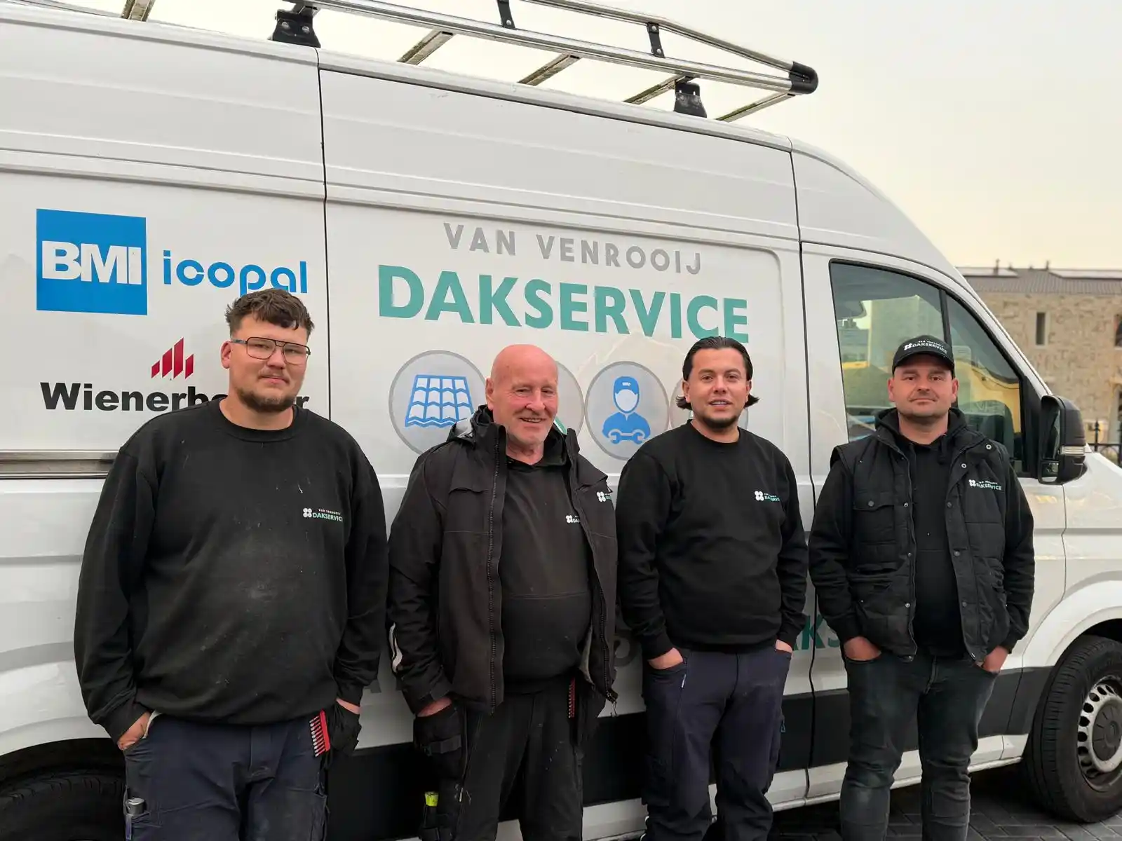 Vier dakdekkers in werkkleding poseren voor een bestelbus met Van Venrooij Dakservice logo en dakbedekkingsmerken.
