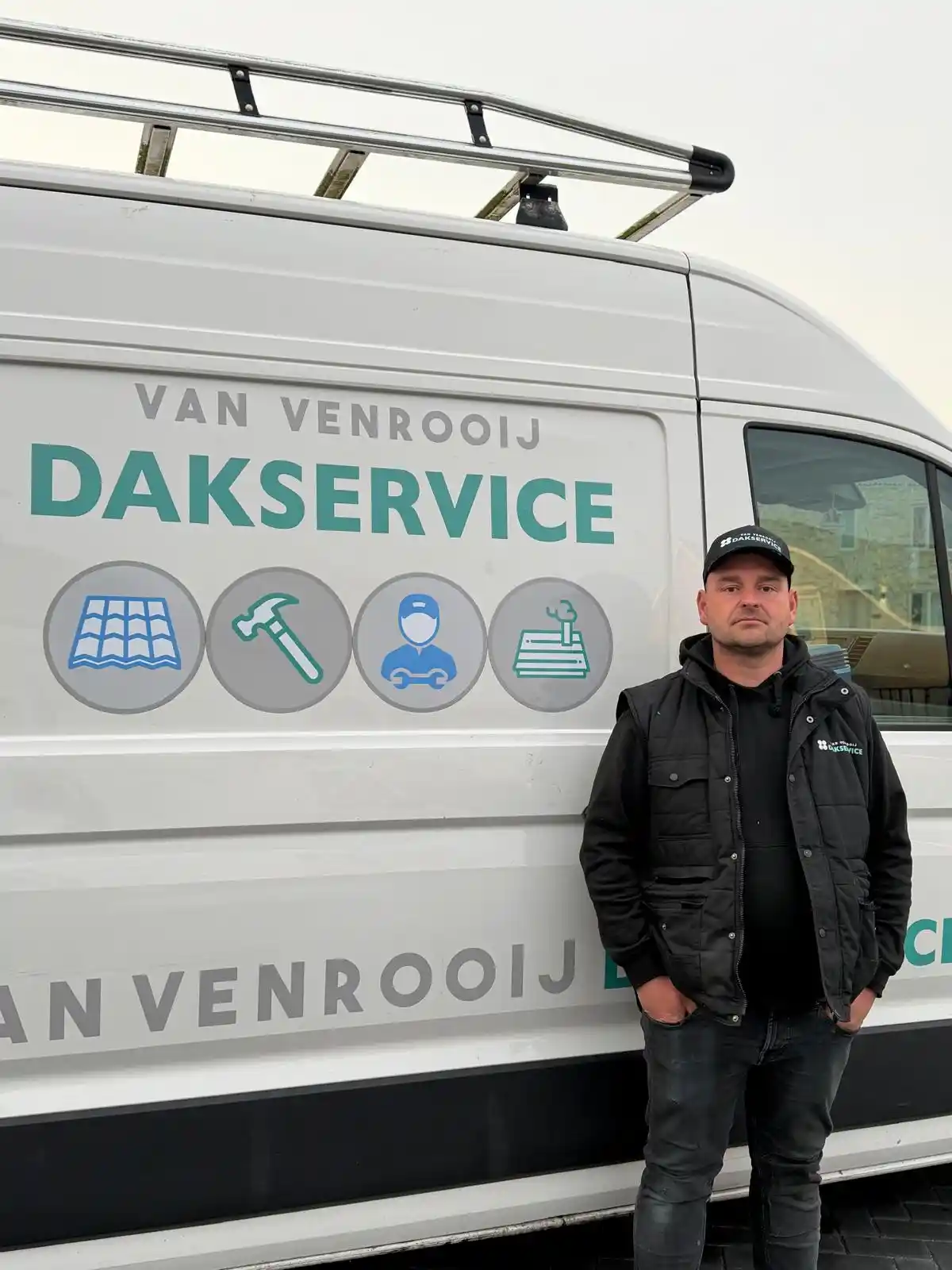 Dakdekker in zwarte jas en pet met Van Venrooij Dakservice-logo staat naast bedrijfsbus met dakservice-tekst en pictogrammen.