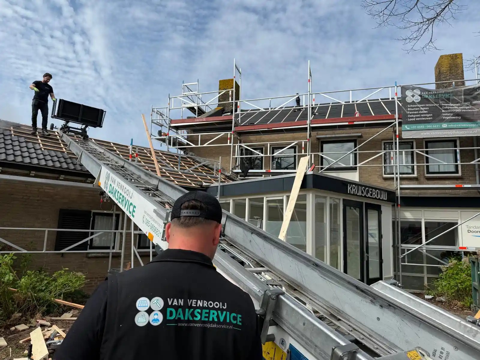 Dakdekker met Van Venrooij Dakservice shirt bedient hijskraan voor dakpannen op steiger bij renovatie van pannendak.